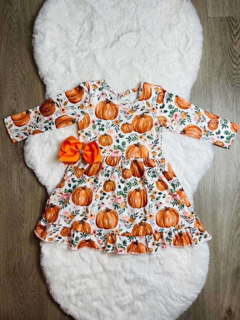 May include: A white dress with a pumpkin and floral print, featuring long sleeves and a ruffled hem. An orange bow is attached at the waist. The dress is designed for children and is suitable for fall or Halloween.