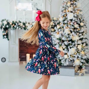 May include: A long-sleeved navy blue dress featuring a repeating holiday print of nutcrackers, ballerinas, rocking horses, and Christmas trees. The dress has a fitted bodice and a flared skirt. A bright red hair bow is worn in the hair.