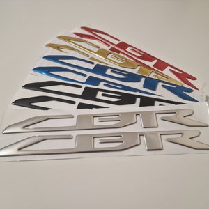 May include: A set of six "CBR" car emblems in different colors, including silver, black, blue, gold, and red. The emblems are on white backing paper.