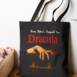 Könnte beinhalten: Schwarze Tragetasche mit dem Text "Bran Stoker's Original Dracula" in Rot und einer Hand, die aus einem Sarg greift. Die Tasche hat schwarze Griffe und einen goldfarbenen Sarggriff.