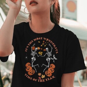 May include: Black t-shirt with a Halloween design. The graphic features dancing skeletons, pumpkins, and fall leaves, with the text "IT'S THE MOST WONDERFUL TIME OF THE YEAR".