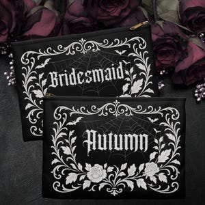 May include: Two black zipped pouches with white gothic text and decorative floral and spiderweb designs. One reads "Bridesmaid," the other "Autumn." The pouches have gold zips and are set against a dark background with roses.