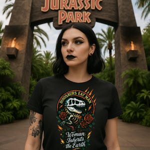 May include: Black t-shirt with a dinosaur skeleton graphic, red roses, and the text "DINOSAURS EAT MAN" and "Woman Inherits the Earth". The background features the Jurassic Park sign and lush greenery.