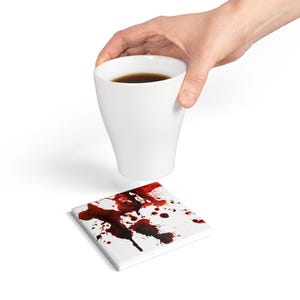 Puede incluir: Posavasos de cerámica blanca con diseño de salpicaduras de sangre roja. Una taza de cerámica blanca con café flota sobre el posavasos.