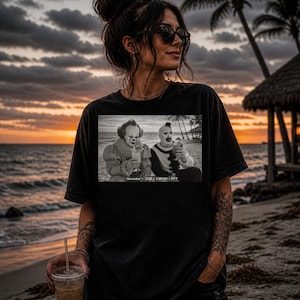 May include: Black t-shirt with a monochrome image of two clowns on a beach, holding drinks. The image includes the text "November 1, 2026 | 5:00 PM | 67°F". The background shows a sunset over the sea.