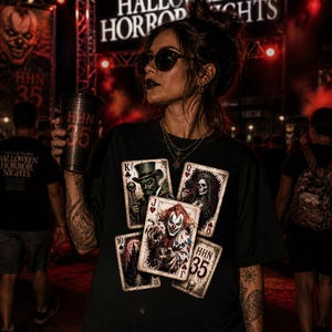 Könnte beinhalten: Schwarzes T-Shirt mit Spielkartenmotiven mit Horror-Illustrationen. Die Karten zeigen einen Clown, ein Skelett und andere gruselige Charaktere. Das Shirt trägt auch den Text "HHN 35". Ein schwarzer Becher mit "HHN 35" wird im Bild gehalten.