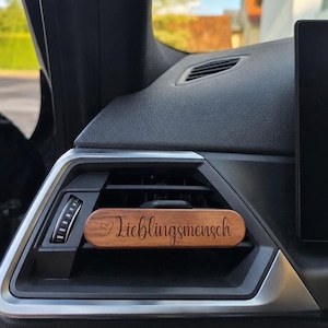 Könnte beinhalten: Ein rechteckiger Auto-Duftbaum-Clip aus Holz, der an einer Lüftungsdüse befestigt ist. Das Holzstück hat eine dunkelbraune Oberfläche mit dem eingravierten Wort "Lieblingsmensch" und einem kleinen Herzsymbol links neben dem Text.