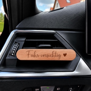 Könnte beinhalten: Ein rechteckiger Auto-Duftbaum-Clip aus Holz, der an einer schwarzen Lüftungsdüse befestigt ist. Das hellbraune Holz trägt den eingravierten deutschen Schriftzug "Fahr vorsichtig" in Schreibschrift, gefolgt von einem kleinen Herzsymbol rechts.