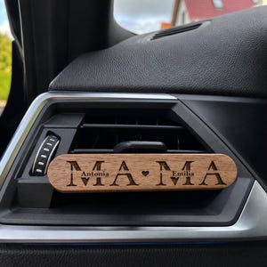 Könnte beinhalten: Ein rechteckiger Auto-Lufterfrischer-Clip aus Holz, der an einer Lüftungsdüse befestigt ist. Das Holz ist mit dem lasergravierten Schriftzug MAMA versehen, wobei die Namen Antonia und Emilia ein kleines Herzsymbol zwischen den M-Buchstaben flankieren.