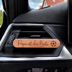 Könnte beinhalten: Ein rechteckiger Auto-Lüftungsclip aus Holz mit der eingravierten deutschen Aufschrift 'Papa ist der Beste' in Schreibschrift, ergänzt durch eine kleine Fußballgrafik auf der rechten Seite, befestigt an einer schwarzen Kunststoff-Lüftungsdüse.