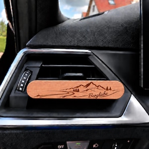 Könnte beinhalten: Ein rechteckiger Auto-Lufterfrischer-Clip aus Holz, der an einer Lüftungsdüse im Armaturenbrett befestigt ist. Das hellbraune Holz zeigt eine lasergravierte Berglandschaft und den Schriftzug "Bergliebe" in einer kursiven Schriftart.