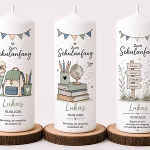 Könnte beinhalten: Drei weiße zylindrische Kerzen mit Illustrationen zum Schulanfang, darunter ein Schulranzen, Bücher, ein Globus und ein Wegweiser. Jede Kerze zeigt den Text Zum Schulanfang, den Namen Lukas, das Datum 10.08.2026 und deutsche Motivationssprüche.