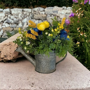 May include: A small, galvanized metal watering can filled with a bouquet of yellow and blue flowers. The flowers are arranged in a rustic, farmhouse style.