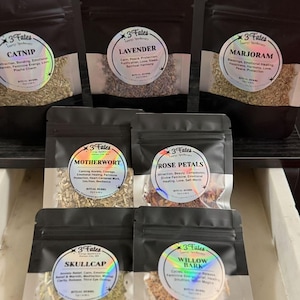 Könnte beinhalten: Eine Auswahl an schwarzen Beuteln, gefüllt mit getrockneten Kräutern, jeweils mit einem holografischen Etikett. Die Etiketten zeigen die Namen "Catnip", "Lavender", "Marjoram", "Motherwort", "Rose Petals", "Skullcap" und "Willow Bark".