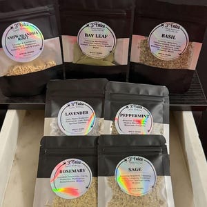 May include: An assortment of dried herbs in black pouches with clear labels. The labels feature the text "3 Fates" and herb names like "Ashwagandha Root," "Bay Leaf," and "Basil." The pouches are arranged in a drawer, showcasing various herbal products.