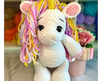 May include: A white crocheted unicorn plush toy with a pink and yellow yarn mane and horn. The unicorn has black eyes and a pink nose.