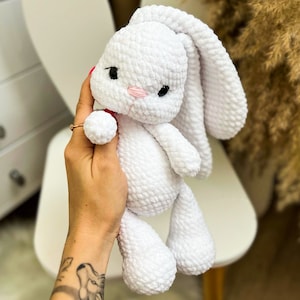 Puede incluir: Un juguete de conejo de crochet blanco con nariz rosa y ojos negros. El conejo está siendo sostenido por una mano.