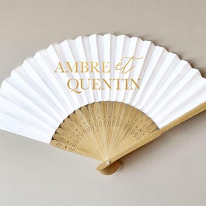 May include: White paper fan with a natural wood handle. The fan is folded and features the gold text "AMBRE et QUENTIN". The fan is open and displayed against a neutral background. Ideal for weddings or special events.