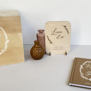 Peut inclure: Un ensemble de livre d'or pour mariage en bois, comprenant une boîte, un panneau et un carnet. La boîte et le carnet portent les noms "Manon & Yann" et la date "18 Mai 2024". Le panneau indique "Livre d'or" et "Laissez-nous un mot doux".