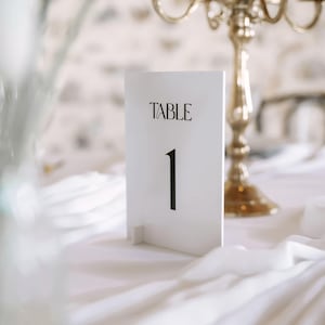 Pode incluir: Um cartão branco com o número da mesa com a palavra «TABLE» em escrita preta acima do número «1». O cartão está sobre uma toalha de mesa branca, com um castiçal dourado ao fundo. Decoração de casamento ou evento.