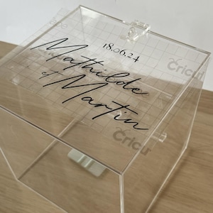 Puede incluir: Caja de acrílico transparente con tapa con bisagras. La parte superior de la caja tiene los nombres "Mathilde et Martin" y la fecha "18.06.24" en escritura negra. La caja está sobre una superficie de madera.