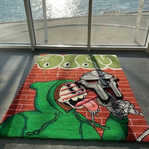 May include: A green and red rug with a cartoon character wearing a green hoodie and a silver mask. The character is holding a pink lollipop and has a long tongue. The rug has a brick pattern background and the words "MF DOOM" are written in green.