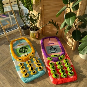 May include: Two colorful, retro-style rugs shaped like cell phones. One is blue and yellow with a globe graphic, the other is pink and purple with a cursor icon. Both have button-shaped number keys.