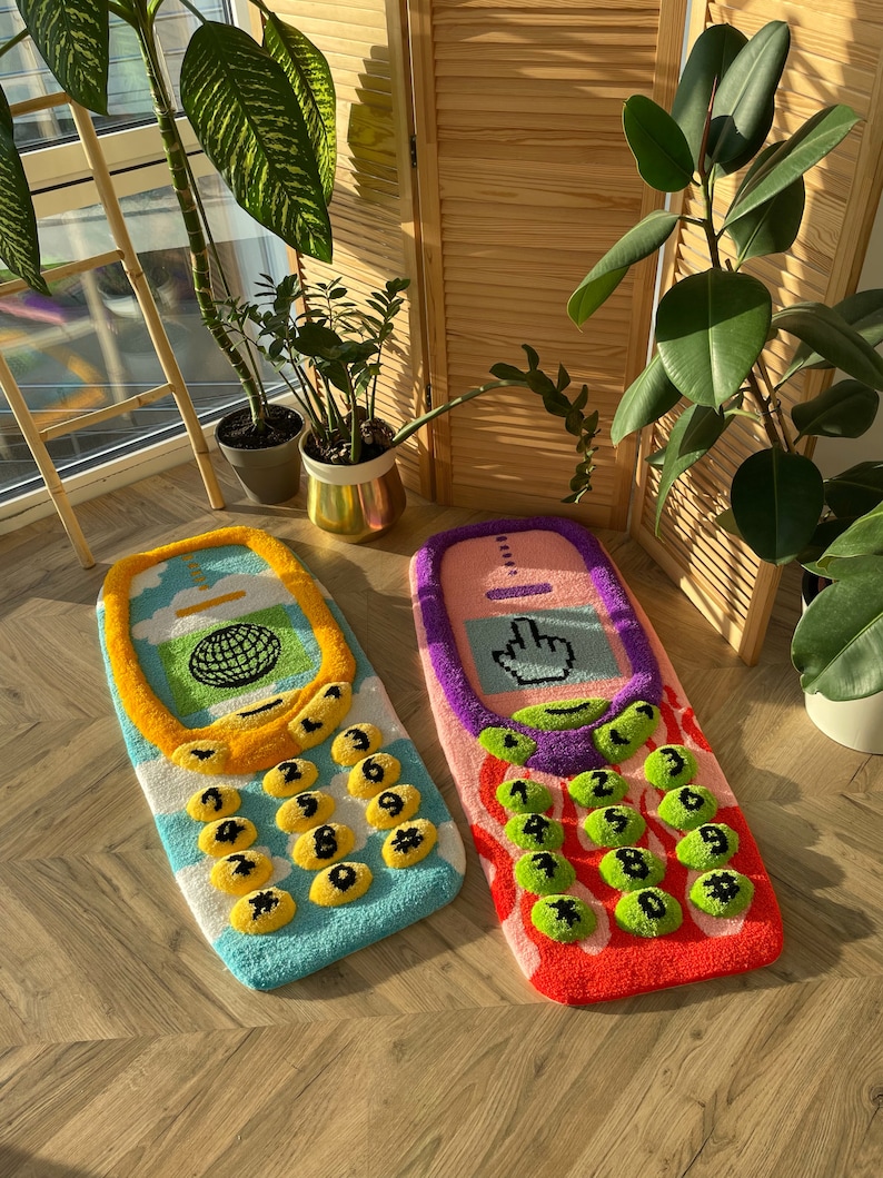 May include: Two colorful, retro-style rugs shaped like cell phones. One is blue and yellow with a globe graphic, the other is pink and purple with a cursor icon. Both have button-shaped number keys.