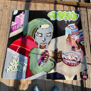 May include: A square, hand-tufted rug featuring a colorful illustration of a masked figure, a bowl of cereal, and graffiti-style text. The rug has a black background with green, red, pink, and gray accents. The text "MF DOOM" is visible.