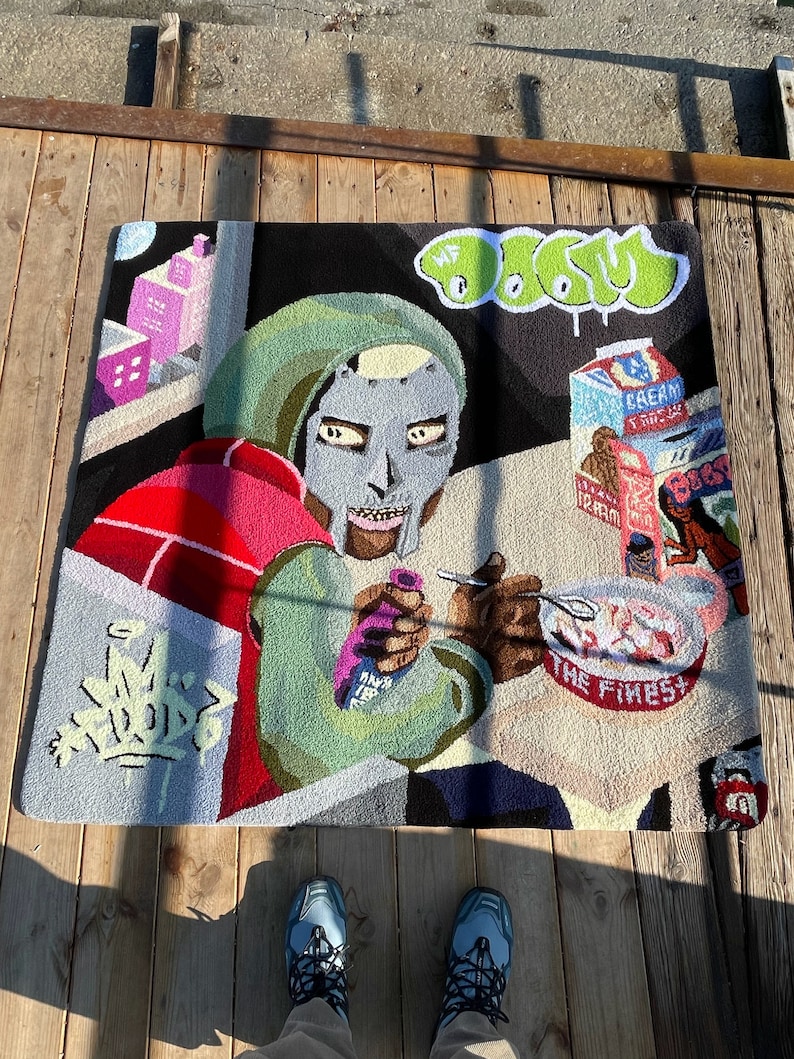 May include: A square, hand-tufted rug featuring a colorful illustration of a masked figure, a bowl of cereal, and graffiti-style text. The rug has a black background with green, red, pink, and gray accents. The text "MF DOOM" is visible.