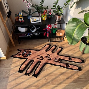 May include: A brown rug in the shape of a cartoon cat with black outlines and details. The rug is on a wooden floor. A black metal shelf with plants and toys is in the background.