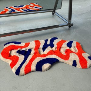 May include: A uniquely shaped rug with a vibrant pattern of orange, blue, and cream. The rug's design features abstract shapes and a soft, textured surface. The rug is placed on a gray concrete floor.