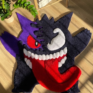 May include: A handmade rug in the shape of a cartoon character, featuring a purple and black body, a red tongue, white teeth, and a red eye. The rug is made of a soft, plush material.