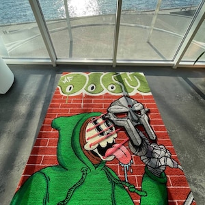 May include: A rectangular rug featuring a cartoon character with a green hoodie and a metal mask, set against a red brick wall background. The rug has a green and white design at the top with the letters "MF". The character's tongue is sticking out.