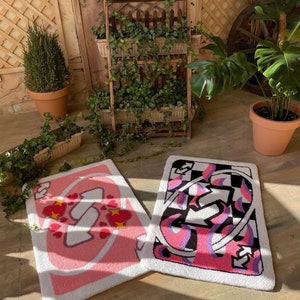 May include: Two rectangular, tufted rugs with card-themed designs. One features a pink and white design with hearts, while the other has a black, white, pink, and purple abstract pattern. The rugs are placed on a concrete surface, with plants and wooden decor in the background.