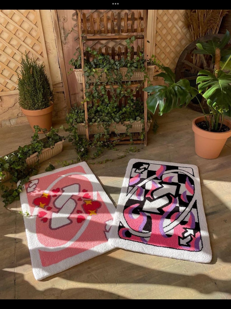 May include: Two rectangular, tufted rugs with card-themed designs. One features a pink and white design with hearts, while the other has a black, white, pink, and purple abstract pattern. The rugs are placed on a concrete surface, with plants and wooden decor in the background.