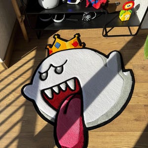 May include: A white, circular-shaped rug featuring a cartoon ghost character with a large open mouth, sharp teeth, and a long pink tongue. The ghost wears a yellow crown with blue and red diamond accents, outlined in thick black borders.
