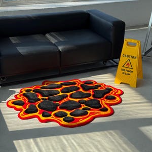 May include: A tufted rug featuring an irregular, organic shape with a design of black rock-like patches surrounded by vibrant red and yellow lava patterns. Next to it stands a yellow plastic caution sign with the text CAUTION THE FLOOR IS LAVA.