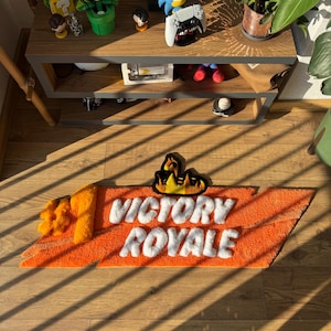 Könnte beinhalten: Ein rechteckiger getufteter Teppich mit einem leuchtend orangefarbenen Hintergrund und einer großen gelben Eins auf der linken Seite. Der Text VICTORY ROYALE ist in Weiß zentriert, mit einer stilisierten gelb-schwarzen Flammengrafik darüber.
