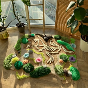May include: A handmade rug featuring a frog skeleton design. The rug has a tan background with a detailed cream-colored skeleton. Green, yellow, and pink tufted accents create a landscape around the skeleton. The rug is placed on a wooden floor.