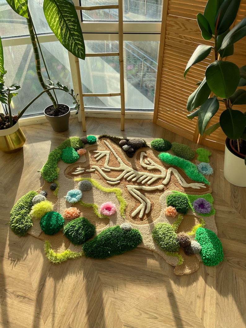 May include: A handmade rug featuring a frog skeleton design. The rug has a tan background with a detailed cream-colored skeleton. Green, yellow, and pink tufted accents create a landscape around the skeleton. The rug is placed on a wooden floor.