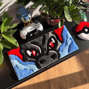 Peut inclure: Un tapis fait à la main représentant le visage d'un dragon en noir, rouge et bleu. Un Pokeball rouge et blanc et une figurine de Sonic le hérisson tenant une manette de jeu blanche sont également visibles. Le tapis est rectangulaire et repose sur une surface noire.