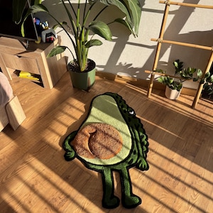 Puede incluir: Una alfombra verde con forma de aguacate y un hueso marrón, con una cara sonriente y piernas. La alfombra está sobre un suelo de madera, con una planta en maceta y una escalera de bambú en el fondo.