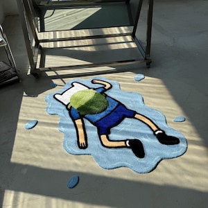May include: A tufted rug shaped like a puddle of light blue water with a cartoon character lying face down in the center. The character wears a blue shirt, blue shorts, black shoes, and a white hat with a green backpack on their back.
