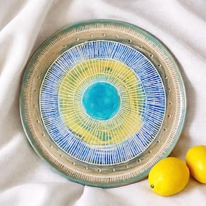 May include: A decorative ceramic plate with a concentric design in shades of blue, yellow, and white. The plate has a turquoise rim and a central blue circle. Two yellow lemons are placed to the right of the plate on a white cloth.