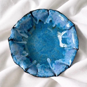 May include: A handmade, ceramic, blue dish with a unique, ruffled edge. The dish features a glossy, textured surface with shades of light and dark blue. The rim is outlined with a dark, contrasting color. The dish is set against a white background.