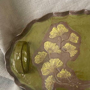 May include: An oval ceramic tray with a glossy olive green glaze and a dark brown rim. The center features a raised, unglazed brown section decorated with a detailed, embossed ginkgo leaf branch pattern in a bright yellow finish.