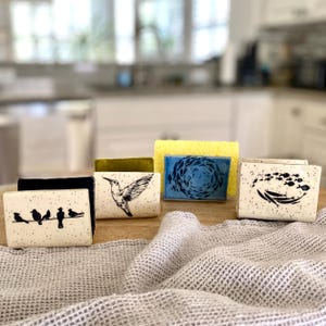 May include: A collection of ceramic sponge holders with various designs. One holder features birds on a wire, another a hummingbird, and a third a school of fish. The holders are speckled with black dots and sit on a wooden surface.