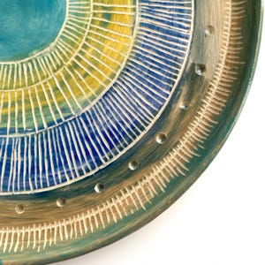 May include: A handmade ceramic plate with a circular design. The plate features concentric rings of blue, yellow, and teal, with white etched lines and silver accents. The plate has a unique, artistic appearance.