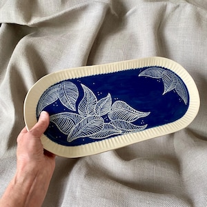 May include: An oval ceramic serving platter with a cream-colored rim and a deep blue center. The blue area features a white leaf design. The rim has a textured, ridged pattern. The platter is held by a hand.